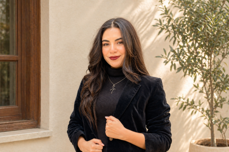 A young woman wearing a black turtleneck and velvet blazer stands confidently in soft natural light against a clean, warm-toned wall with a subtle plant in the background, creating a minimal and elegant look.